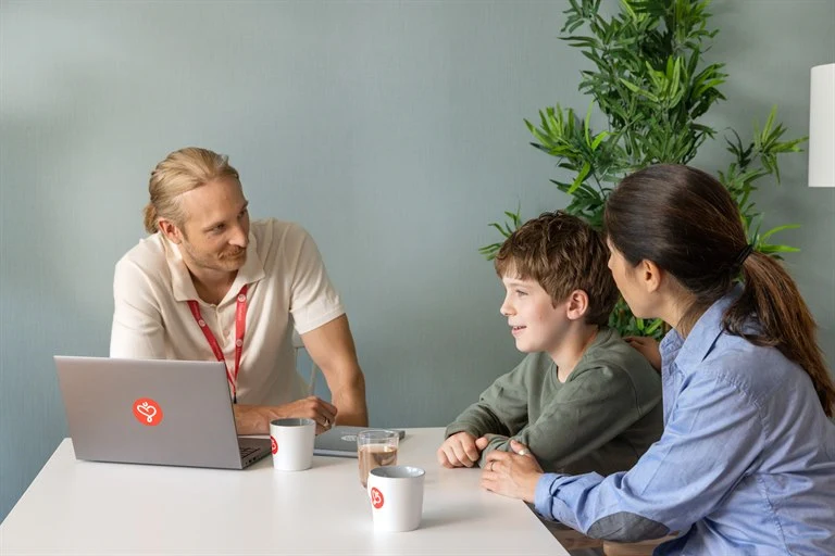 En man en kvinna och en pojke sitter vid ett bord, mannen har en dator och ett nyckelland med Humana logotypen på, de samtalar