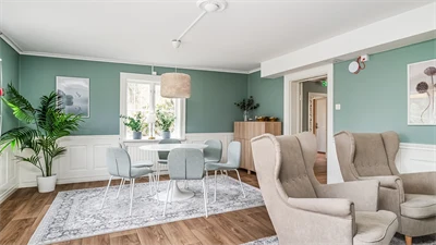 Vardagsrum med gröna väggar och trägolv. I rummet står fåtöljer i beige, ett matbord i vitt med ljusblå stolar runt. 
