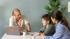 En man en kvinna och en pojke sitter vid ett bord, mannen har en dator och ett nyckelland med Humana logotypen på, de samtalar