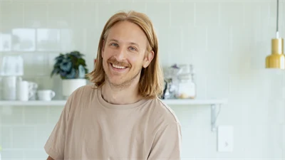 En man med axellångt blont hår, står i ett kök och häller upp kaffe, han är glad