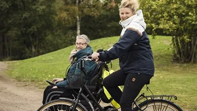 En kvinna åker i permobil och den andra cyklar. Bägge ler mot kameran.