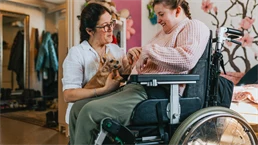 Kvinna i rullstol klappar en liten hund som assistenten håller i