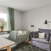 Sovrum målat i vitt med skrivbord i trä, grön stol. Rummer har en säng med grönt överkast och gröna gardiner samt en grå fåtölj. 