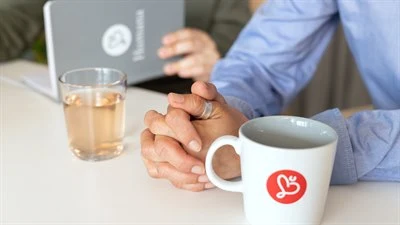 Knutna en händer på ett bord, ett glas med saft och en kaffekopp med Humana logotypen, i bakgrunden är det en person som läser i ett anteckningsblock