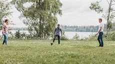 Kolme henkilöä pelaamassa jalkapalloa nurmikolla järven rannalla.