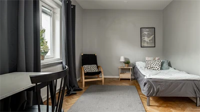 Sovrum med grå matta grå säng mot vit vägg, fönster och skrivbord