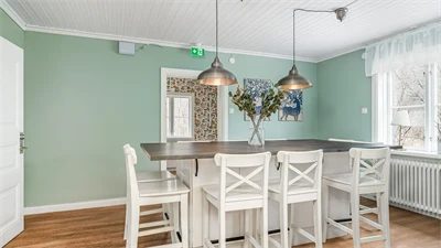 Kök med gröna väggar och trägolv. I rummet står ett matbord med träskiva och stolar i vitt. 