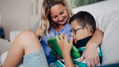 Leende mamma håller om pojke som använder läsplatta i soffa